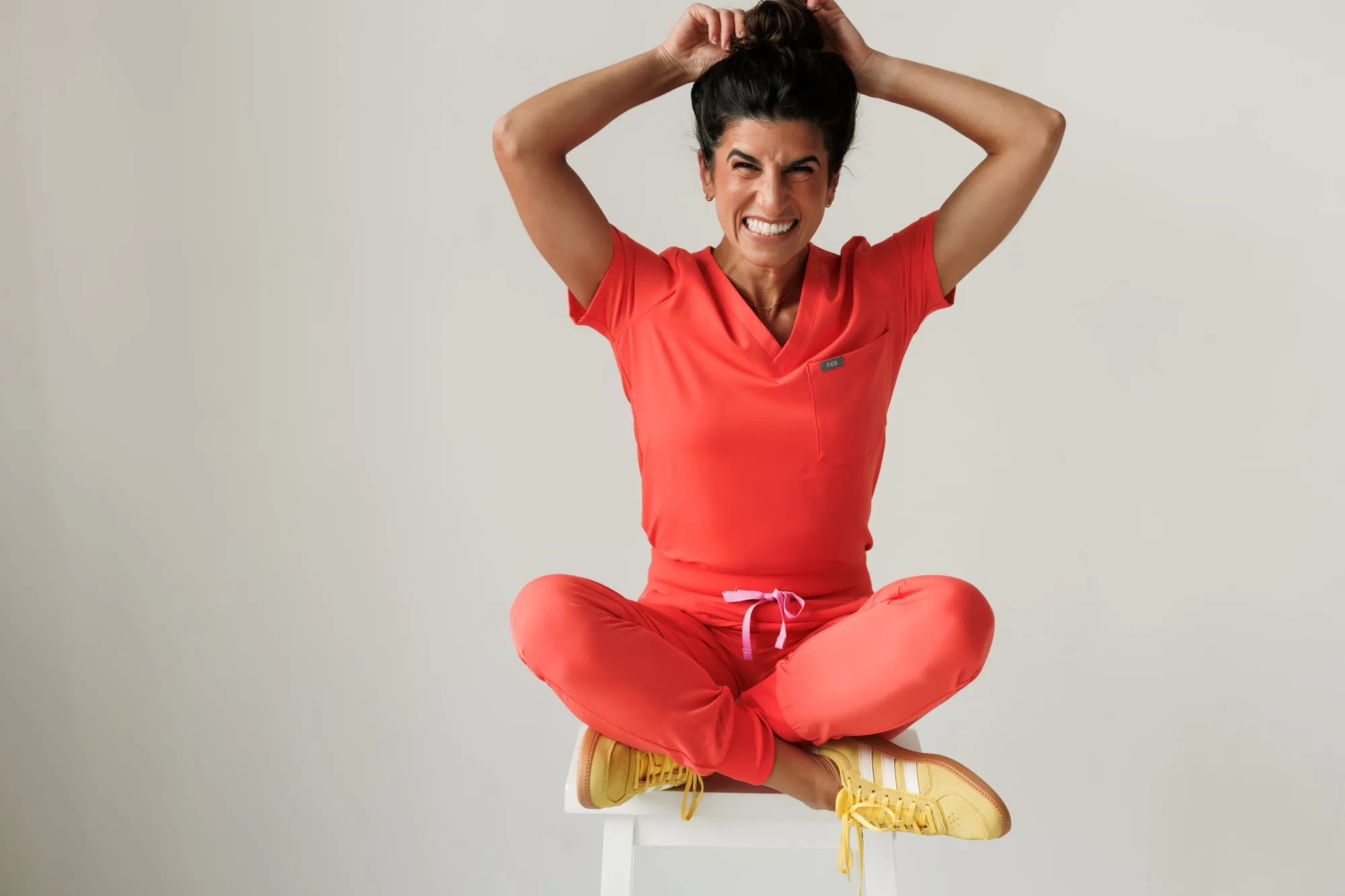 Dr. Caris Miller sitting cross-legged on a stool in coral scrubs and yellow sneakers, hands on her head, grinning