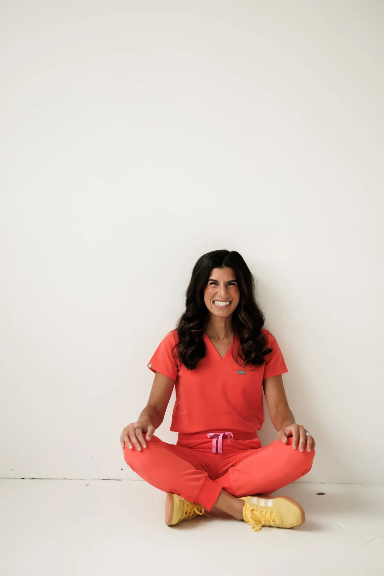 Dr. Caris Miller sitting cross-legged on the floor in coral scrubs and yellow sneakers, smiling