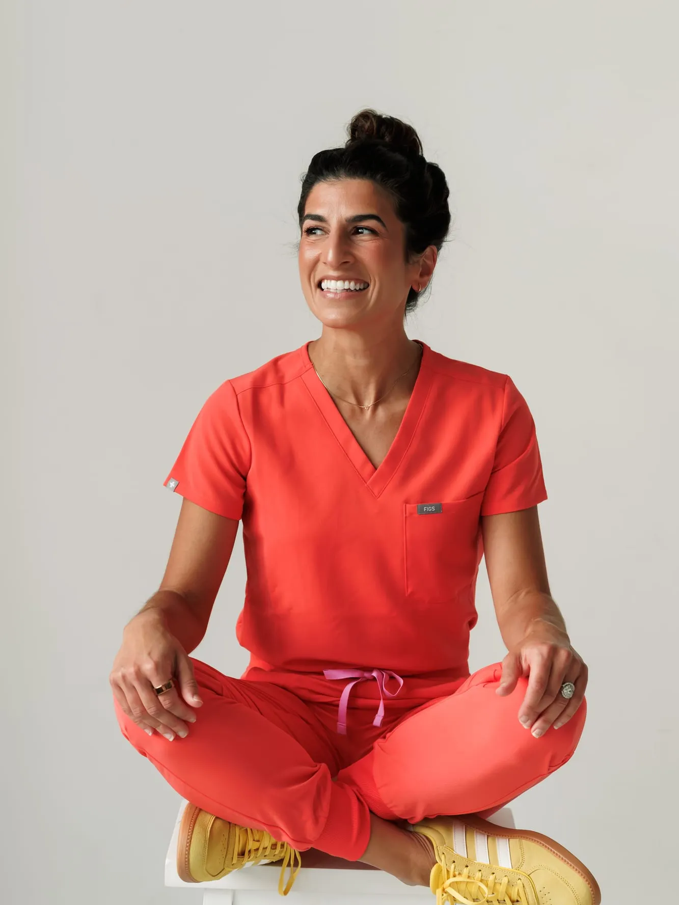 Dr. Caris Miller sitting cross-legged on a stool in coral scrubs and yellow sneakers, smiling